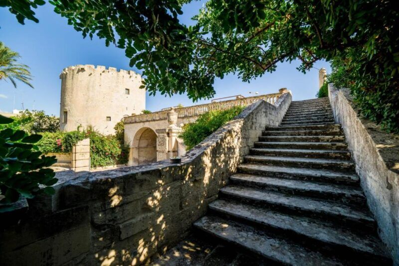 Stone staircase leads up to a garden terrace with an old round tower in the background, surrounded by greenery under a clear blue sky at Torre del Parco, Lecce.