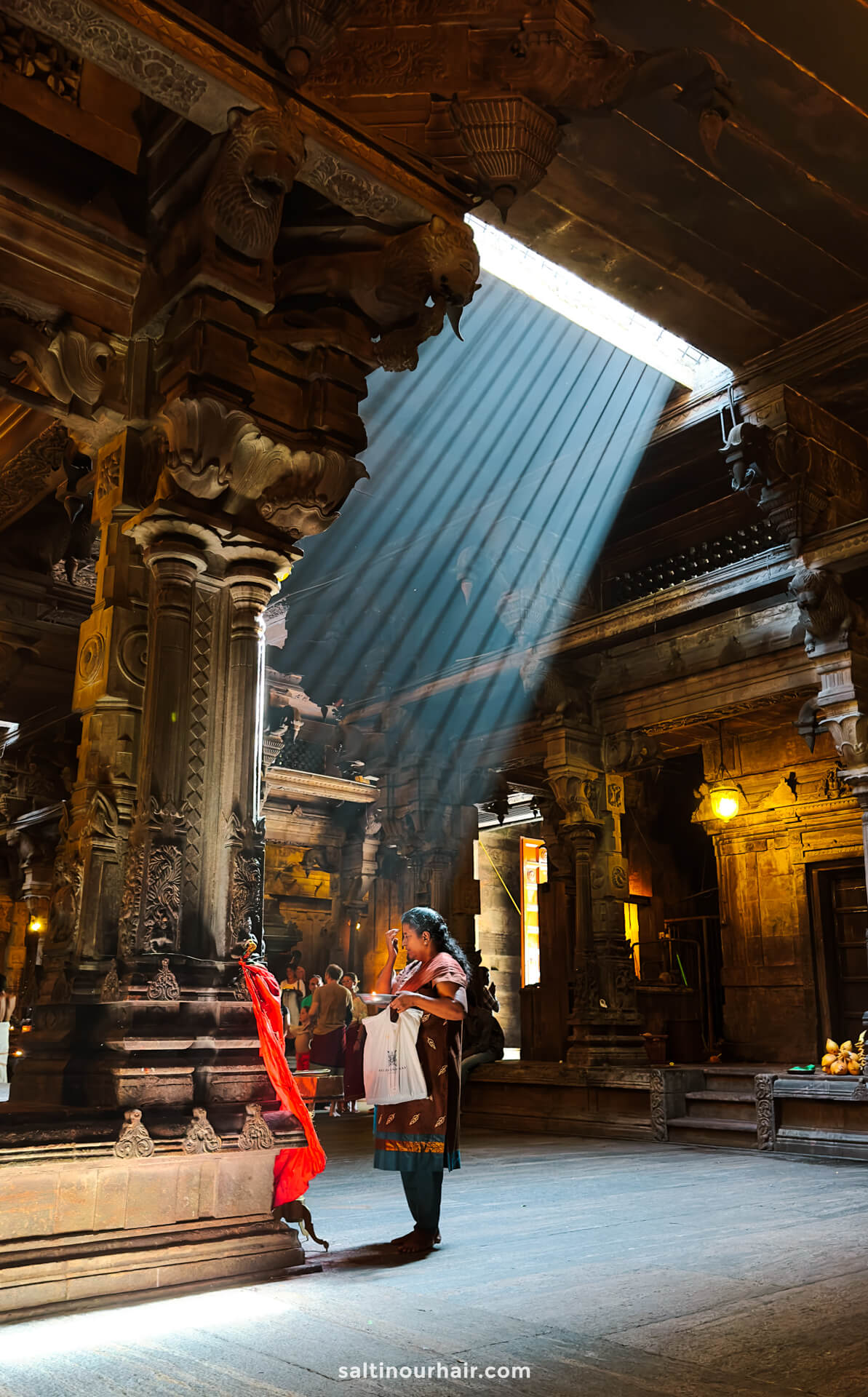 A woman stands in prayer inside Sri Ponnambalavaneswarar Kovil, illuminated by sunlight streaming through an opening in the ceiling.