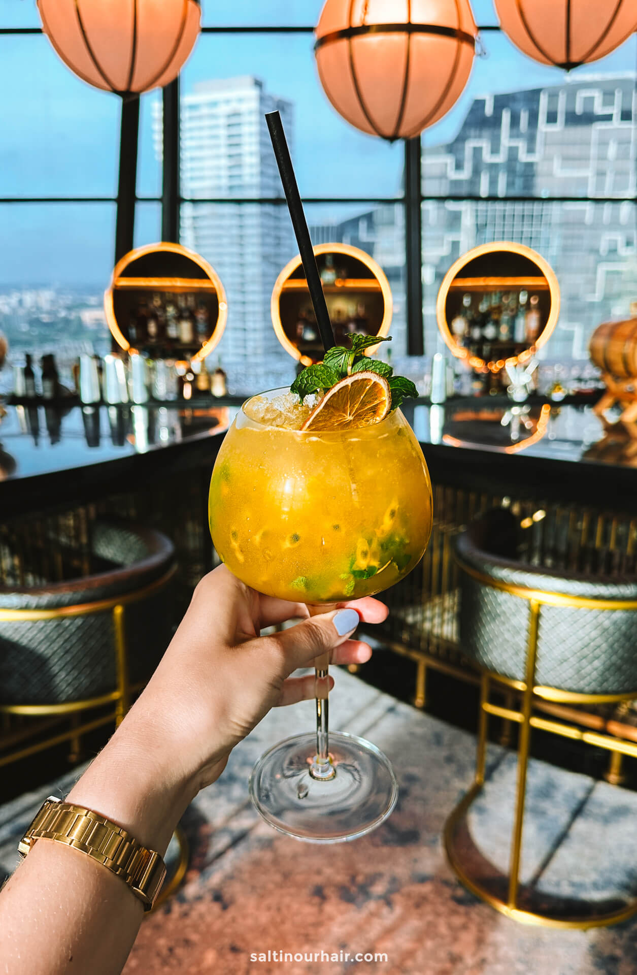 A hand holding a large yellow cocktail with mint and an orange slice garnish at a modern rooftop bar with city views over Colombo, Sri Lanka.