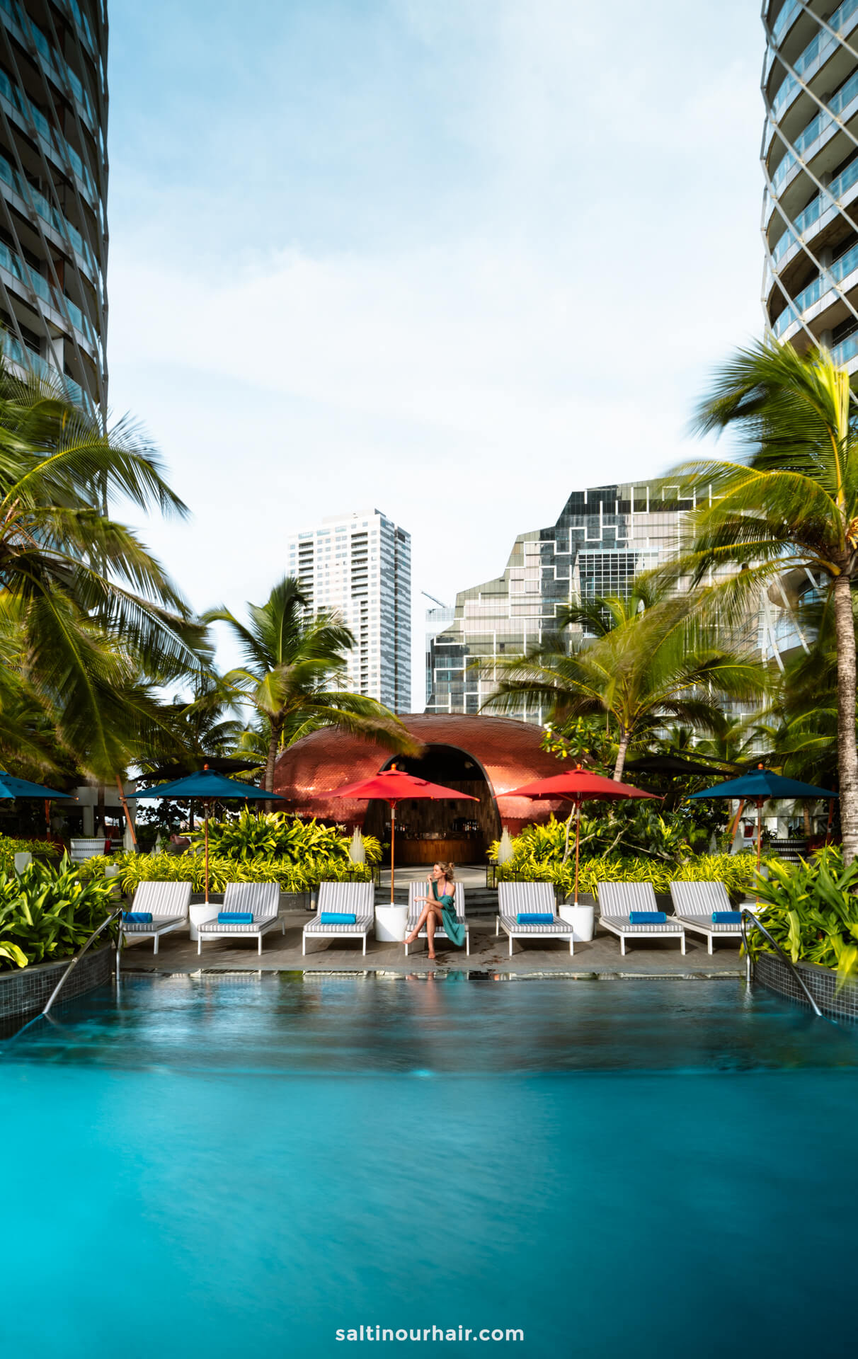 A woman sits on a poolside lounge chair surrounded by palm trees, with tall modern buildings in the background. One of the options of where to stay in Colombo, Sri Lanka.