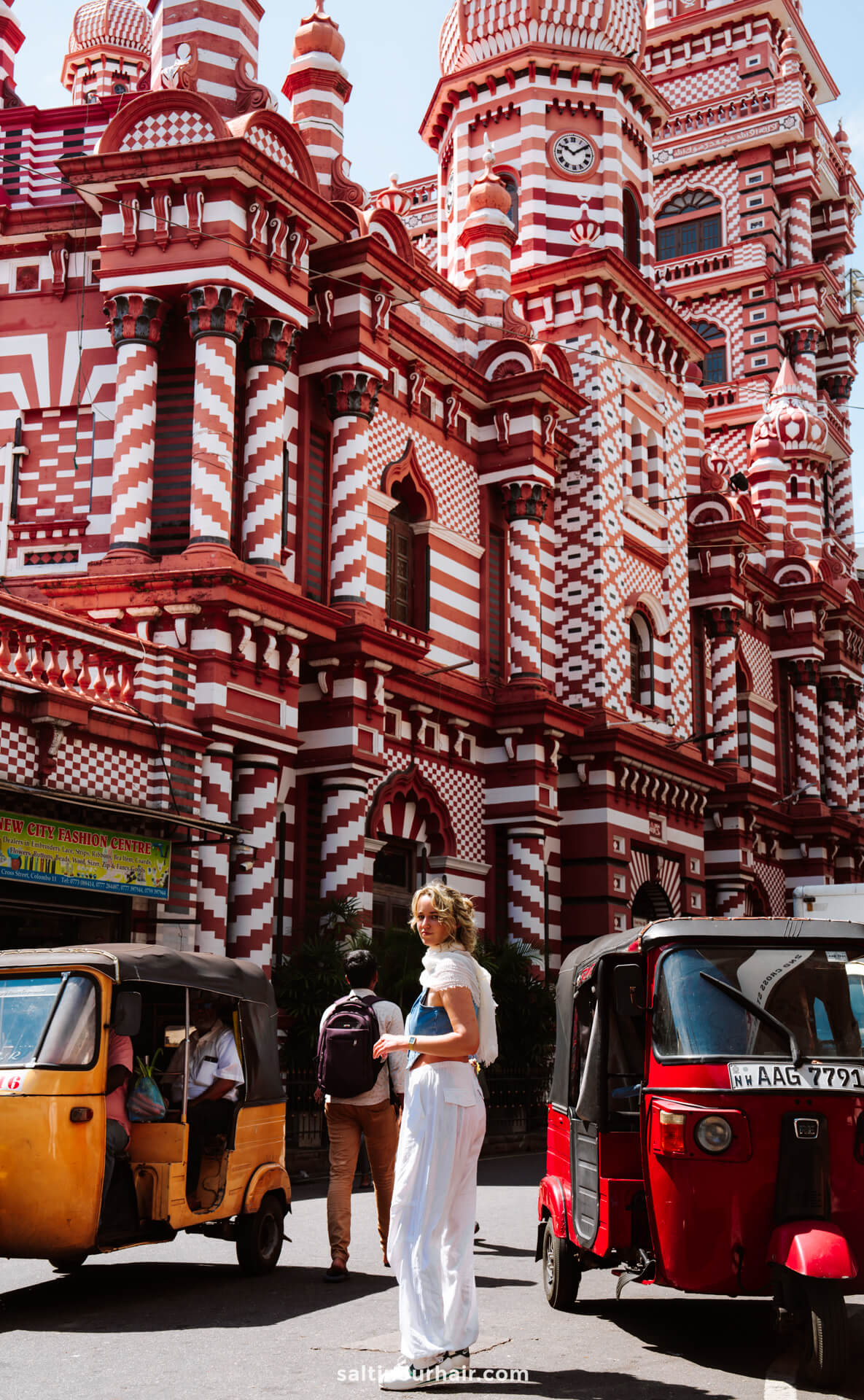A woman in white clothing stands near colorful tuk-tuks in front of the red-and-white patterned Jami Ul-Alfar Mosque in Colombo, Sri Lanka.