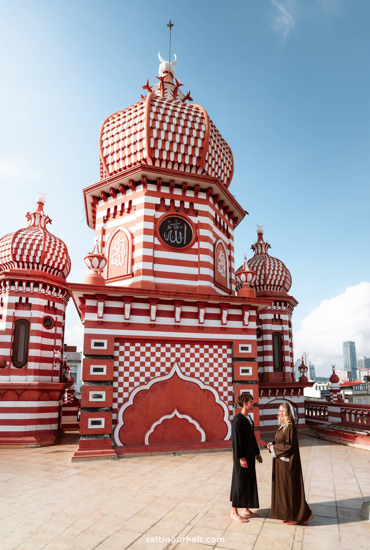 Two people in brown robes stand at the rooftop of the red mosque in Colombo, Sri Lanka with domed roofs and Islamic calligraphy.