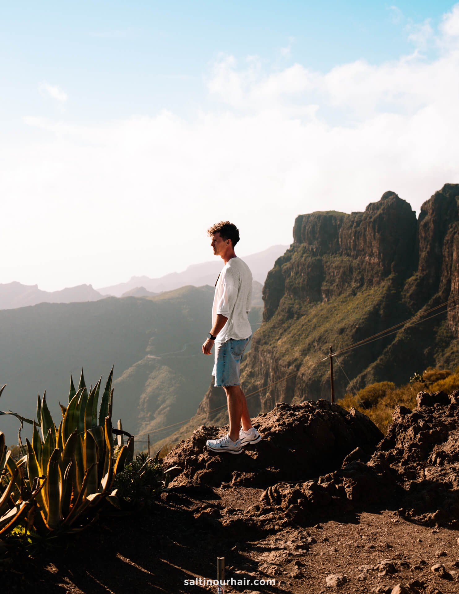 A man in a white shirt and shorts stands on a rocky outcrop, overlooking the stunning mountainous landscape of Masca, Tenerife, under a partly cloudy sky.