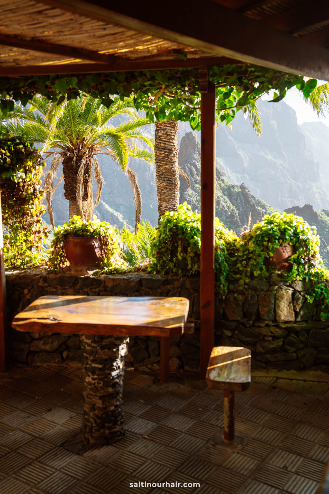Wooden table and stool on a shaded patio with potted plants, stone wall, and palm trees overlooking the sunlit mountainous landscape of Masca, Tenerife.