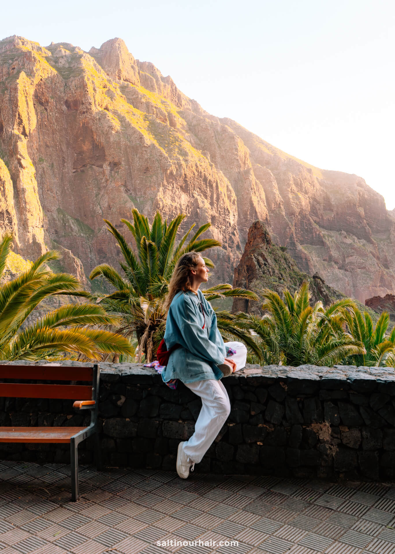 A person wearing a blue shirt and white pants sits on a stone wall, gazing at sunlit mountains and palm trees in the background of Masca, Tenerife.
