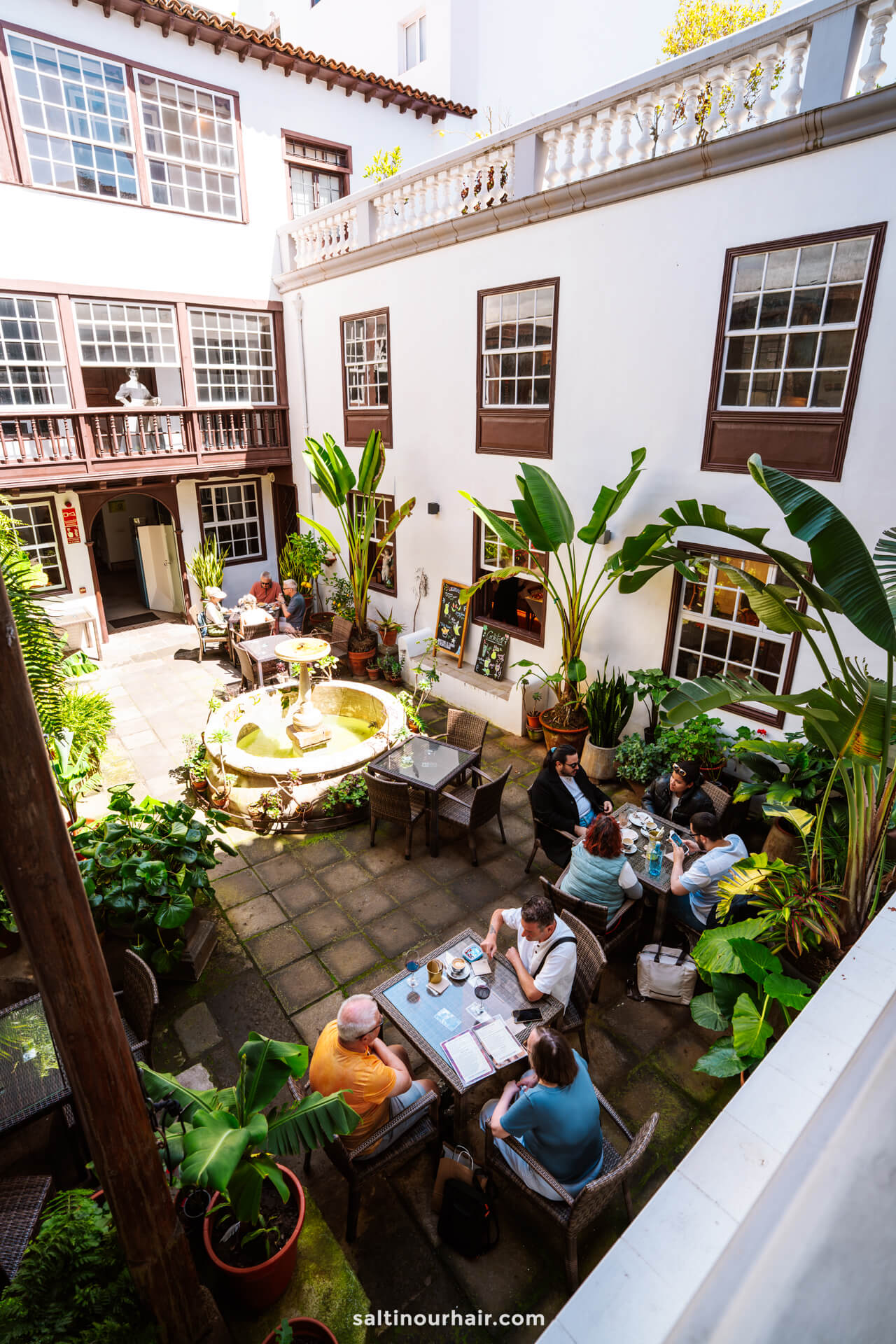 A charming courtyard caf&eacute; inside Casa Museo Cayetano G&oacute;mez in  San Crist&oacute;bal de La Laguna tenerife, with people seated at tables, surrounded by lush plants and a central fountain,.
