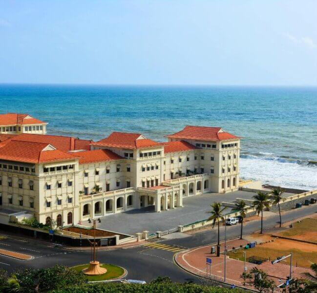 The iconic Galle Face Hotel, a large white colonial-style building with red roofs, sits beside a coastal road, facing the ocean under a clear sky, with palm trees and waves in the background.