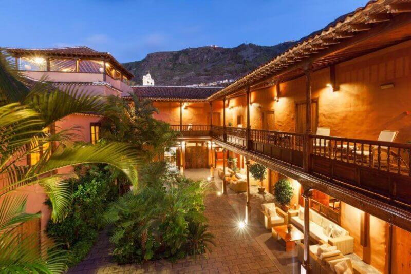 Open courtyard of La Quinta Roja, a traditional Spanish-style hotel in Garachico, with wooden balconies, outdoor seating, lush plants, and warm lighting at dusk, set against a scenic hillside backdrop.
