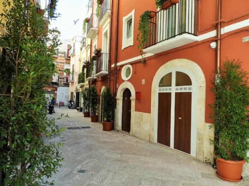 Narrow street with potted plants and red-orange buildings featuring wooden doors and balconies under daylight, near the charming Arco Della Neve in Bari.