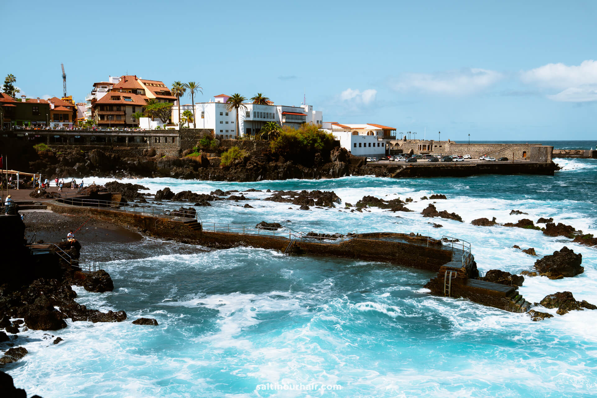 Waves crash against volcanic rocks near Playa San Telmo in Puerto de la Cruz, Tenerife, with buildings and palm trees visible under a clear blue sky.