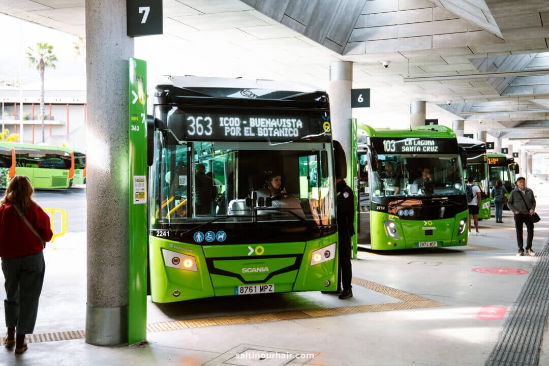 Green city buses are parked at a modern bus terminal in Puerto de la Cruz, with passengers boarding and walking nearby under a concrete roof.