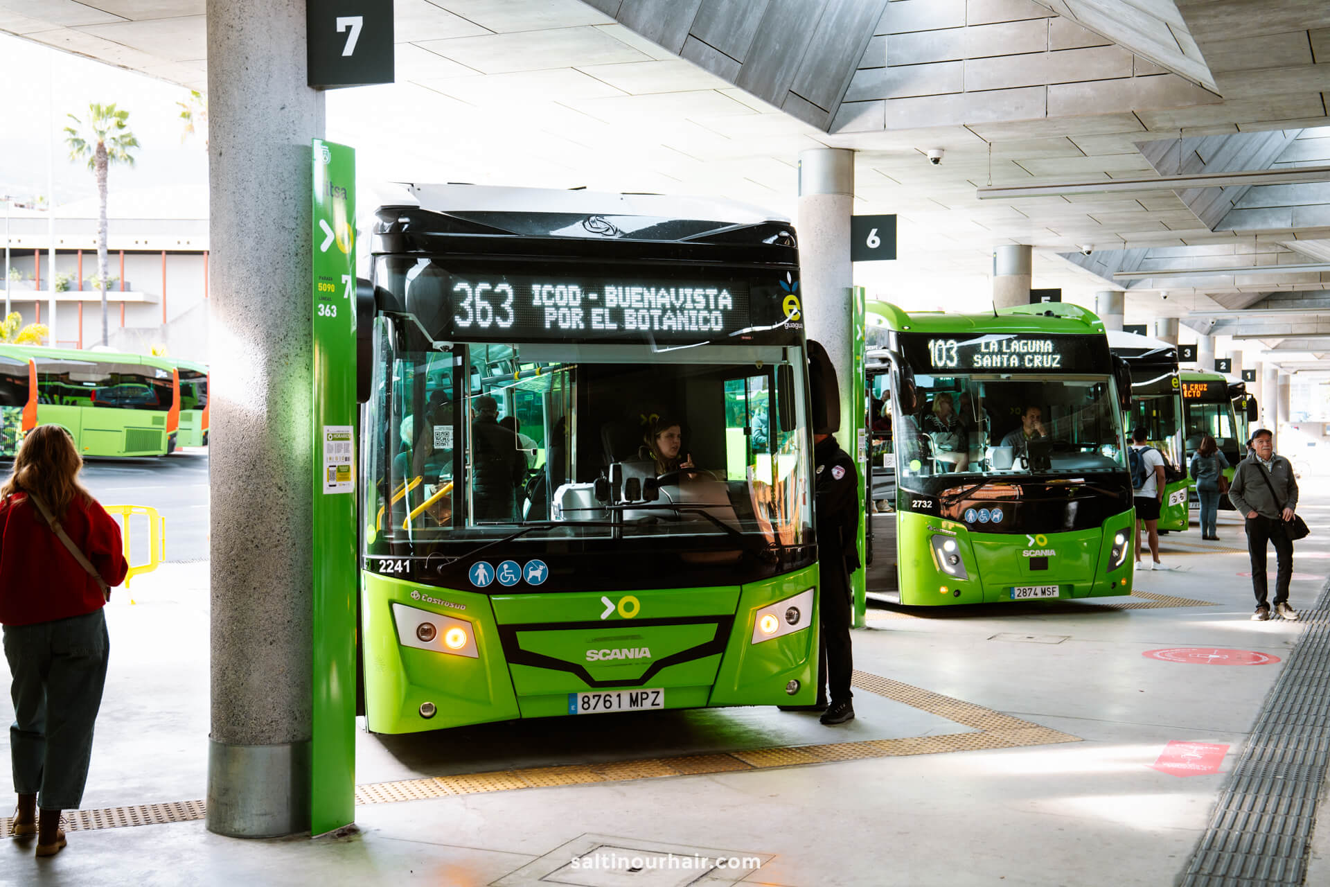 Green city buses are parked at a modern bus terminal in Puerto de la Cruz, with passengers boarding and walking nearby under a concrete roof.