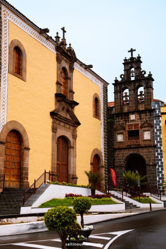 A historic church in la orotava with a yellow facade, arched wooden doors, and an adjacent bell tower adorned with multiple crosses and clocks, set amid landscaped urban surroundings.