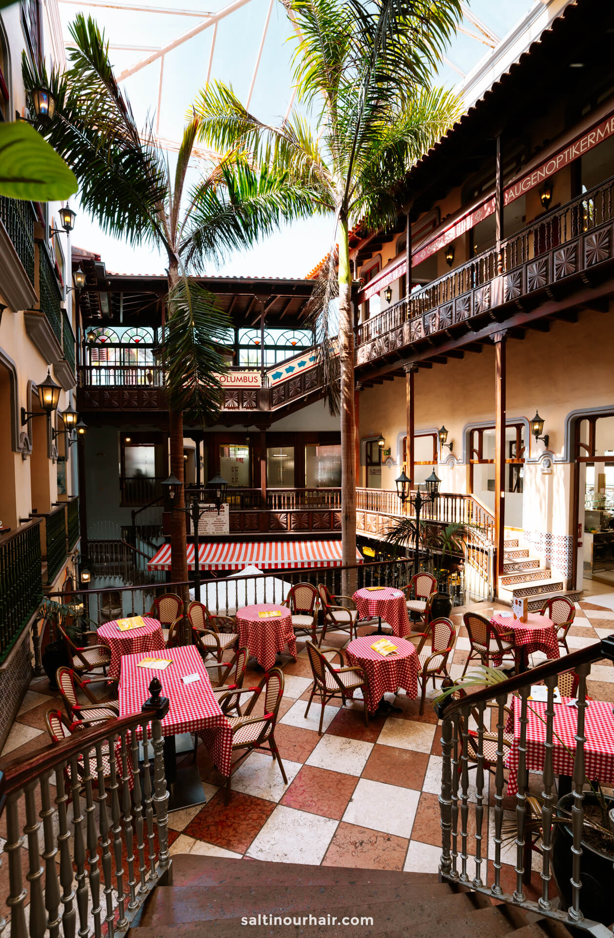 An open-air courtyard in Puerto de la Cruz with red and white checkered tablecloths, wooden chairs, palm trees, and upper-level balconies in a sunlit, Mediterranean-style building.