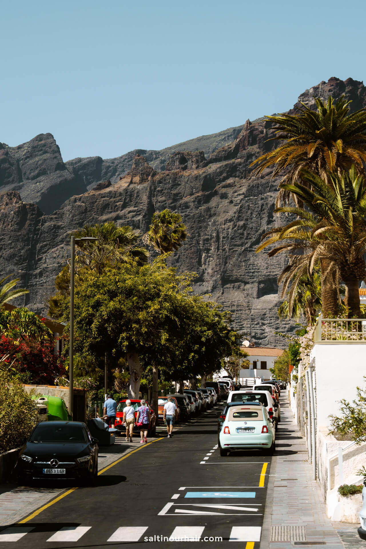 A street in Los Gigantes Tenerife lined with parked cars, palm trees, and pedestrians, with steep rocky cliffs rising majestically in the background under a clear sky.