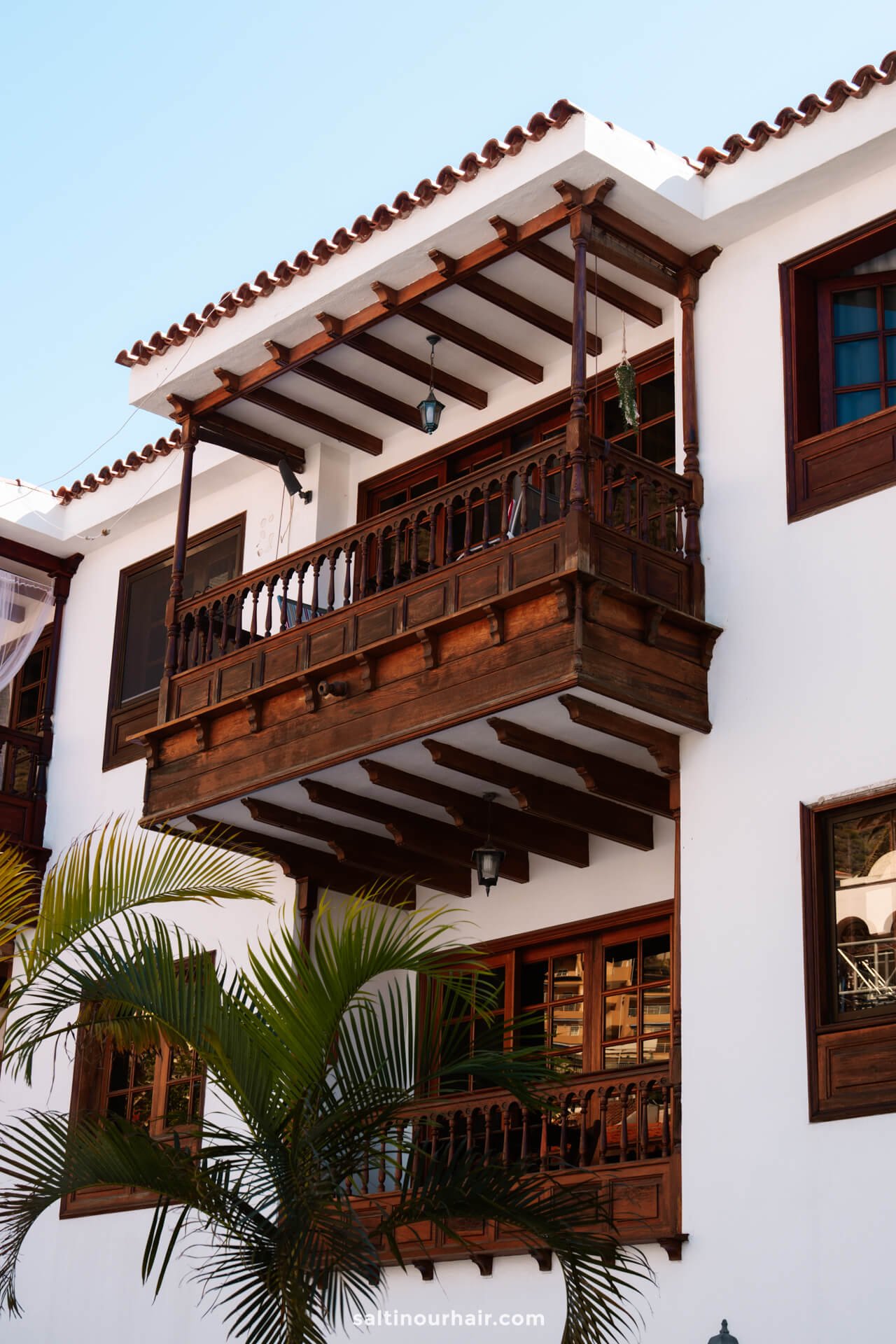 A traditional white building with wooden balconies and windows, featuring a tiled roof and a potted palm in the foreground, captures the charm of Los Gigantes village Tenerife.