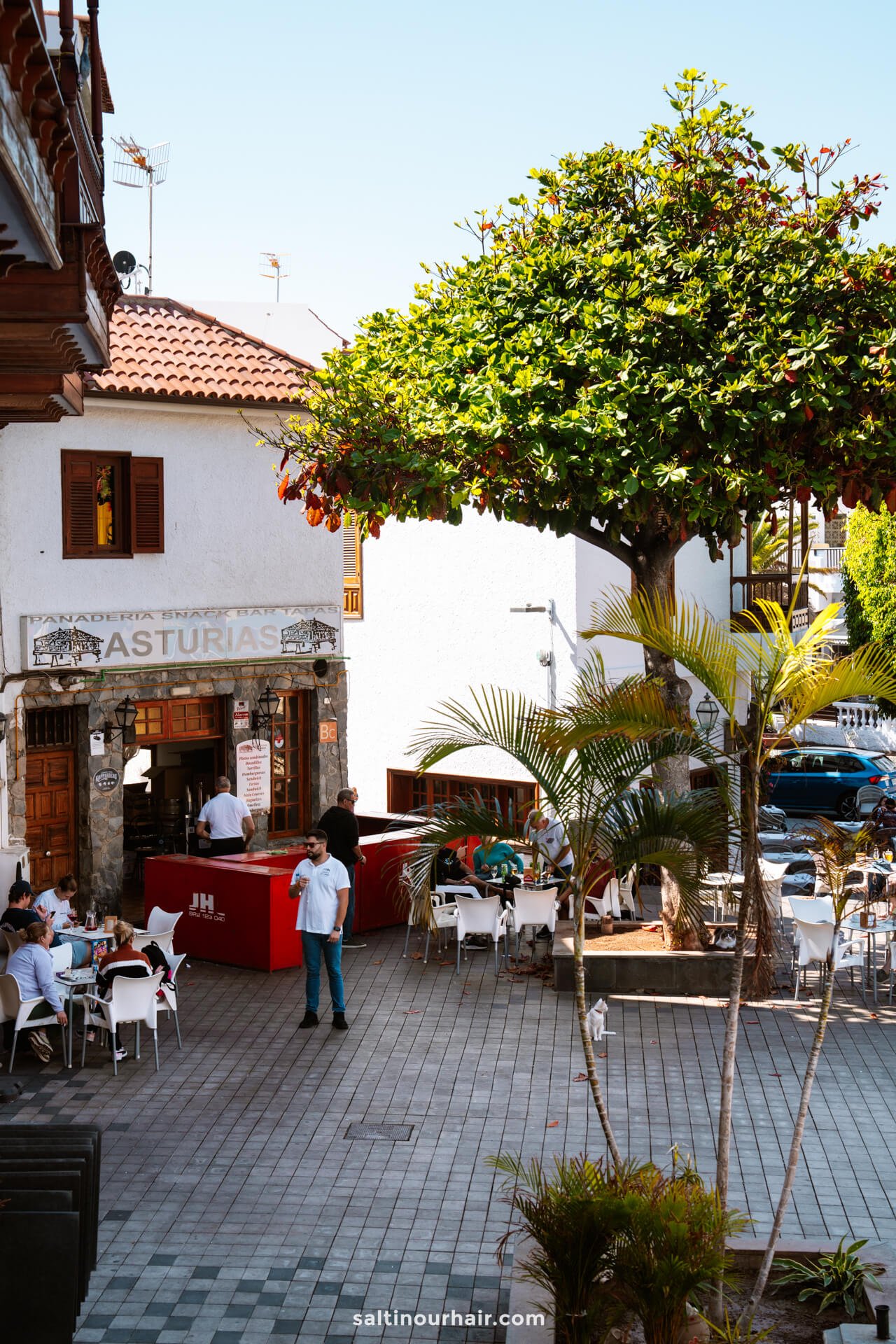 An outdoor cafÃ© with white tables and chairs is situated beside a tree near Los Gigantes Tenerife. People are seated, and a person stands by a red counter outside a building labeled Asturias.