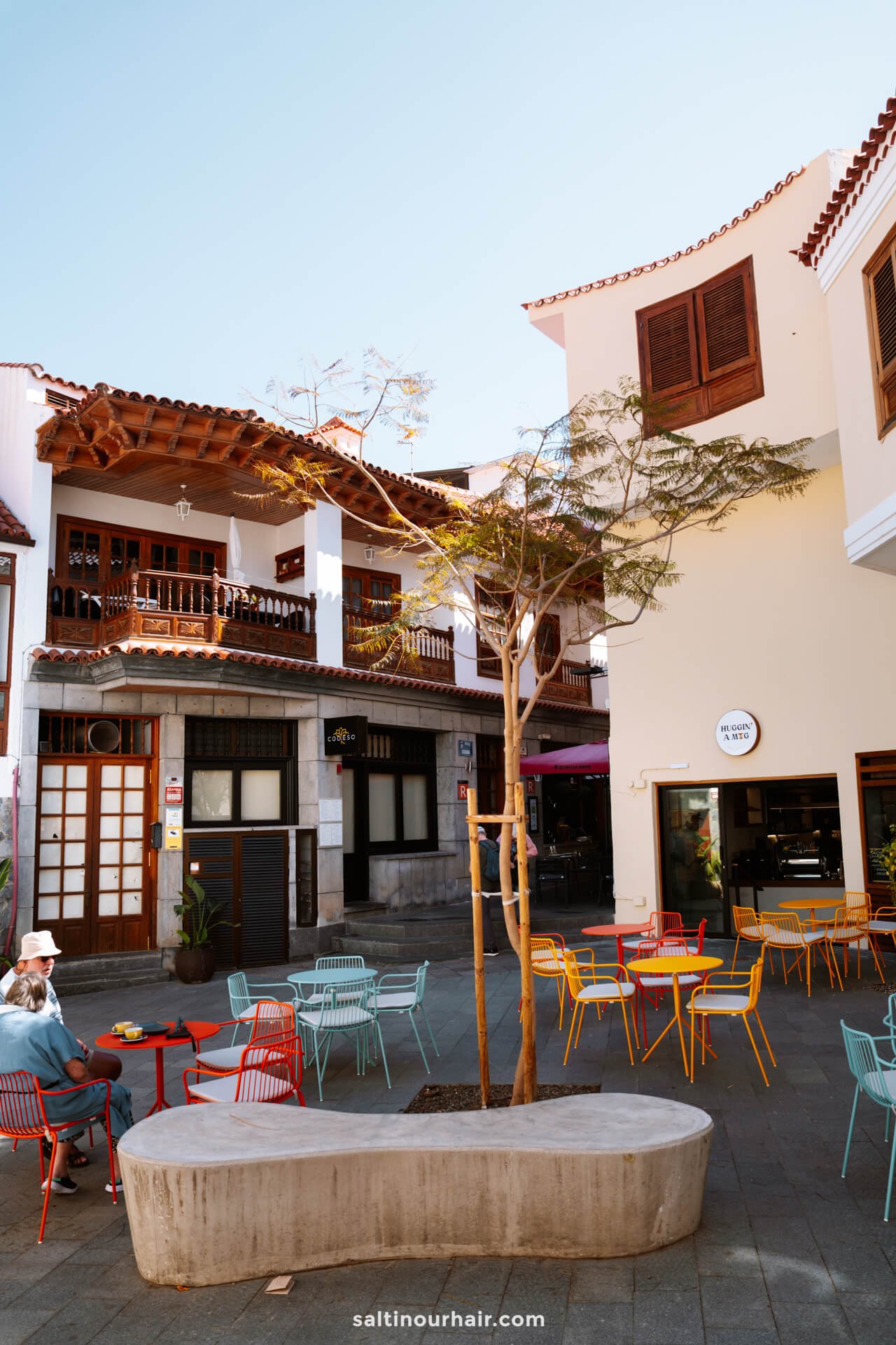 Outdoor cafÃ© scene with colorful chairs and tables, a curved concrete bench, and a tree in a town square bordered by white and yellow buildings with balconies, capturing the charming ambiance of Los Gigantes village Tenerife.