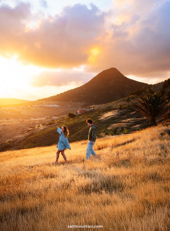 Two people walk through a field of golden grass at sunset in Porto Santo, Madeira, with a mountain and scattered houses in the background.