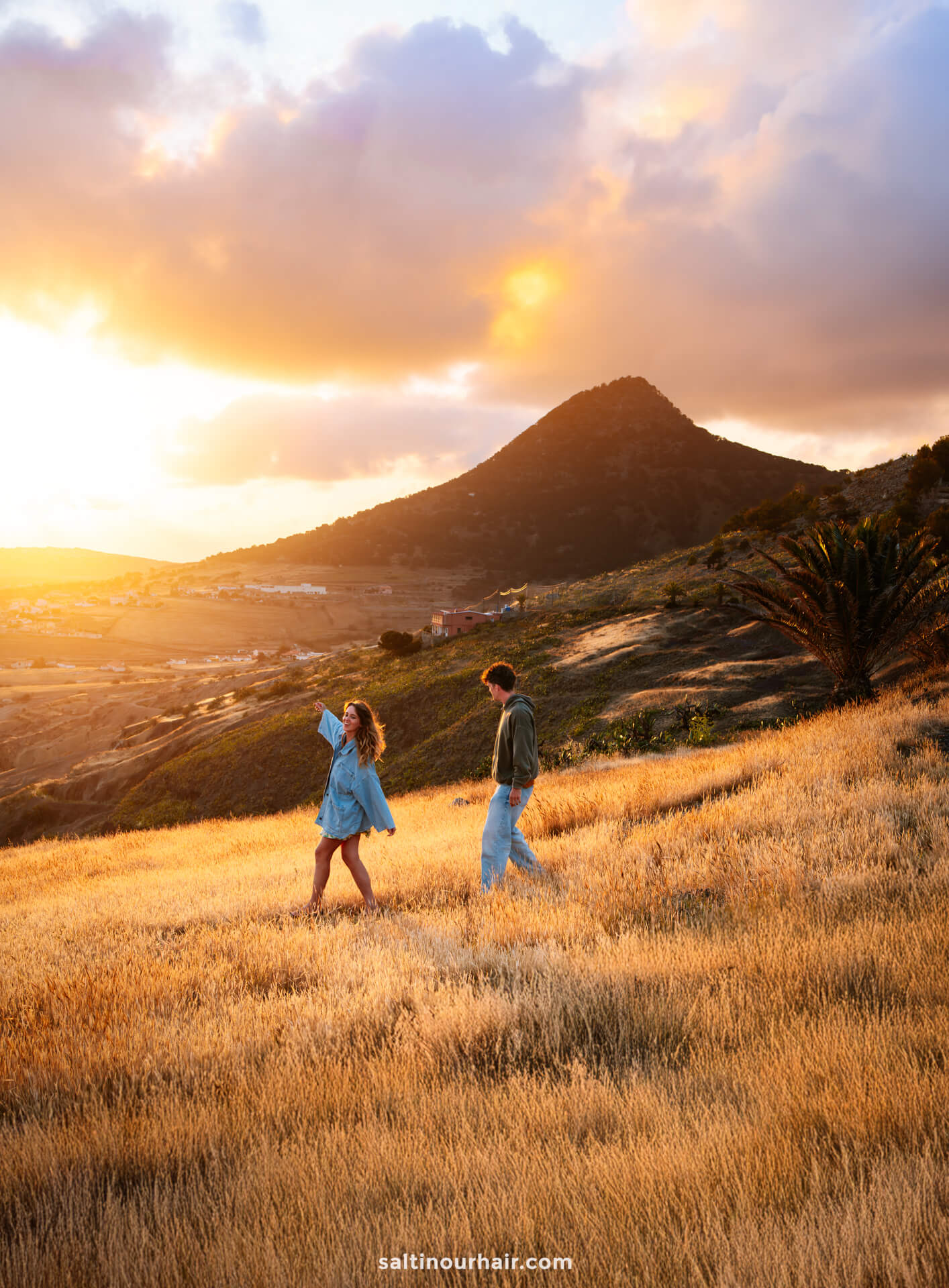 Two people walk through a golden field at sunset on Porto Santo, with a mountain in the background and dramatic clouds in the skyâ€”one of the most picturesque things to do in Porto Santo.