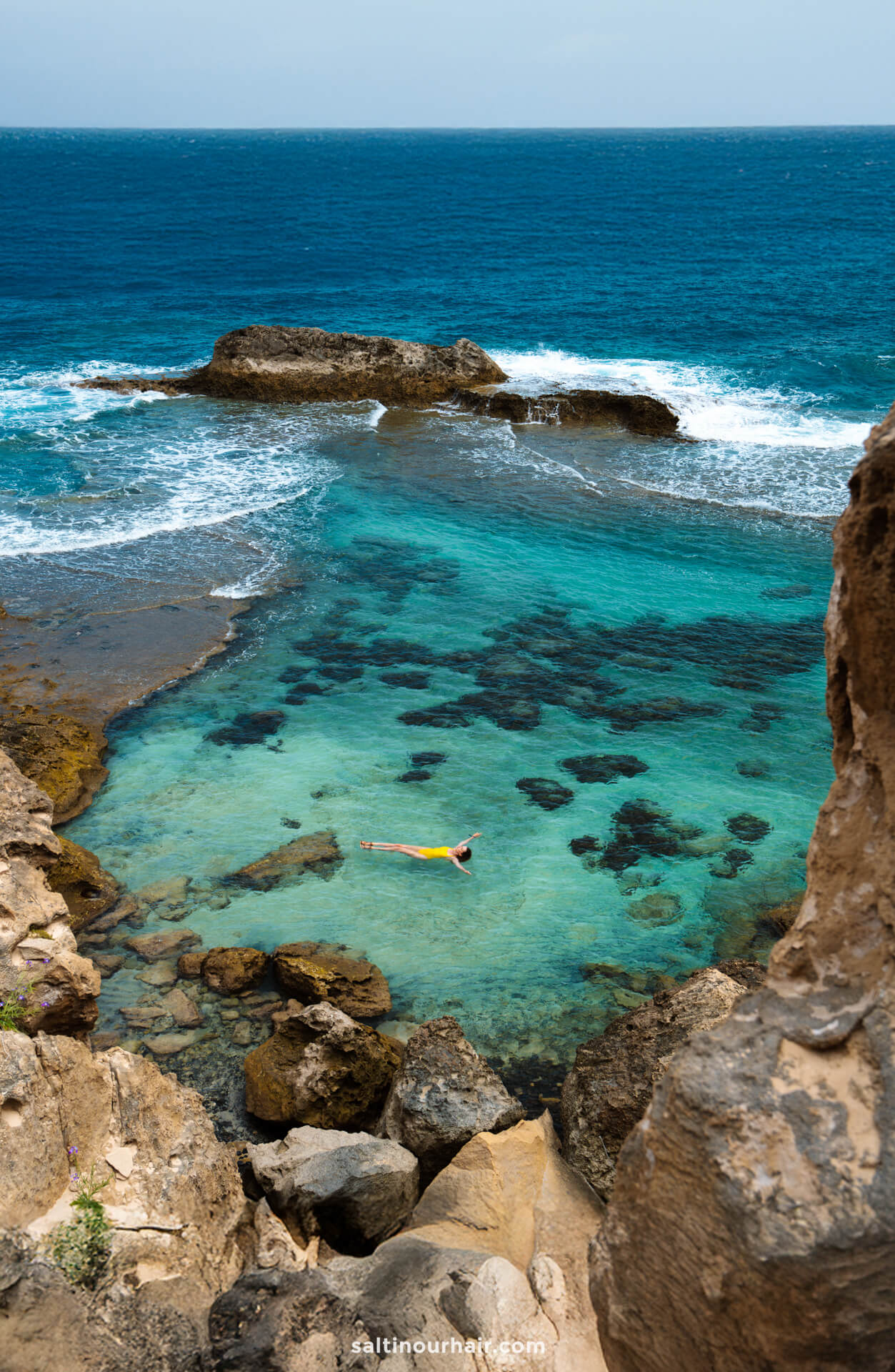 woman floating on their back in a clear turquoise tide pool surrounded by rocks, with waves and the ocean visible in the backgroundâ€”a serene scene and one of the unforgettable things to do in Porto Santo.