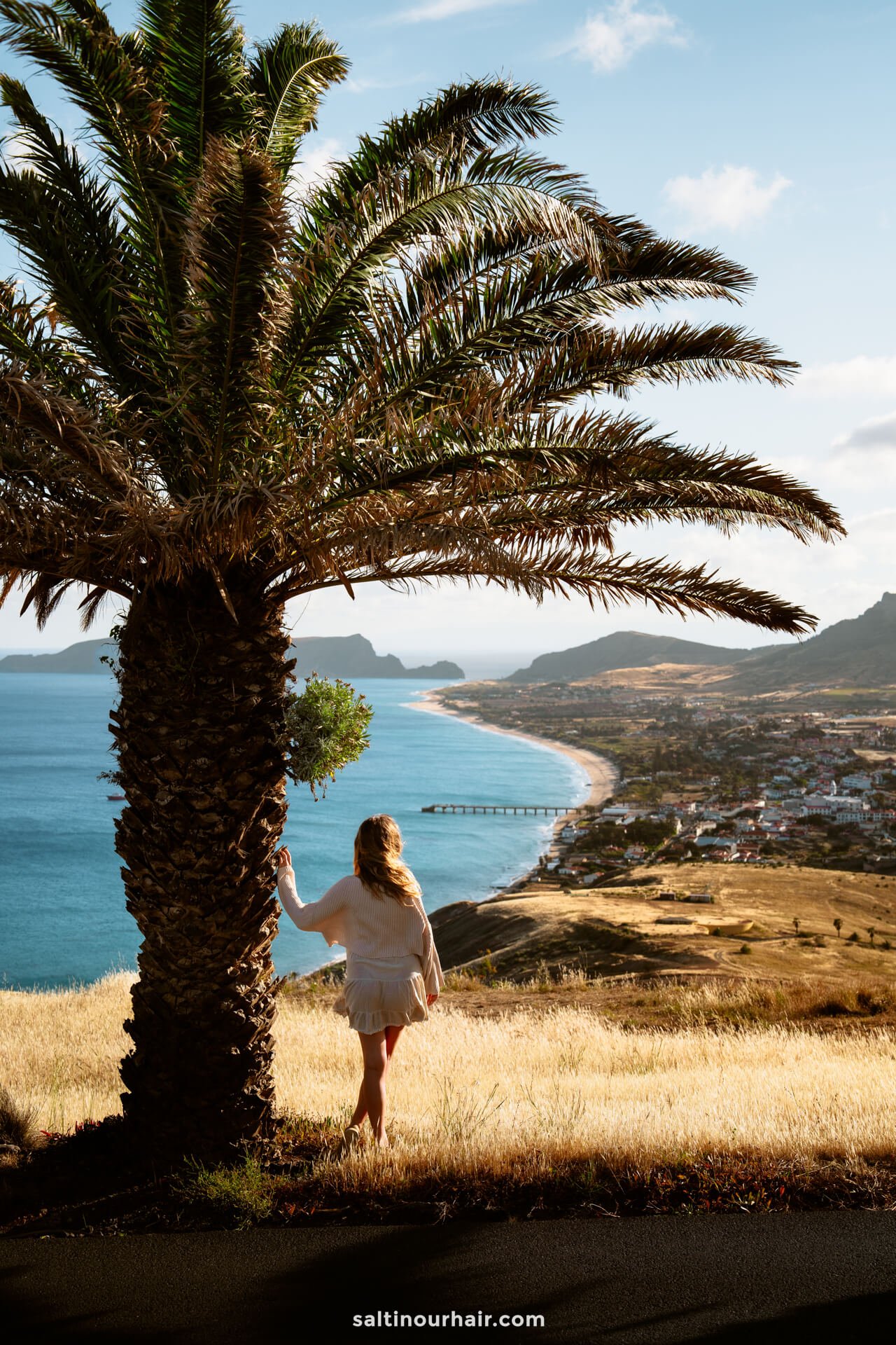 A woman in a white dress stands by a palm tree on a grassy hill, overlooking Porto Santoâ€™s coastal town, golden beach, and ocean with distant mountains beneath a blue skyâ€”one of the stunning things to do in Porto Santo.