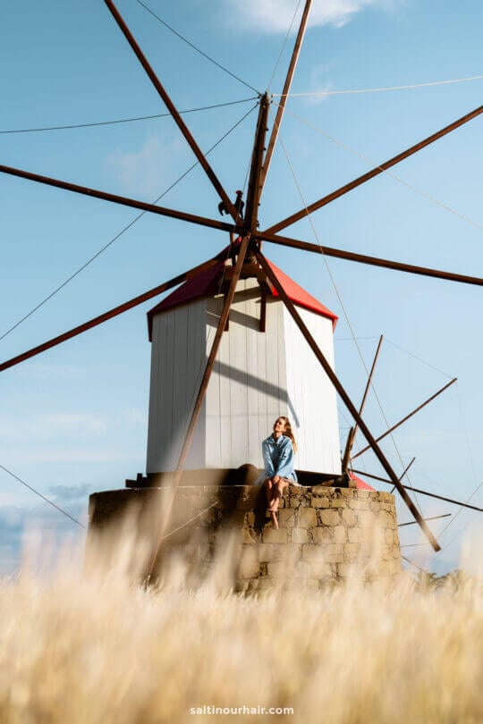 A woman sits on a stone wall in front of a traditional windmill with a red roof and large wooden blades under a clear blue sky, capturing the charm of Porto Santoâ€”a must-see among things to do in Porto Santo.