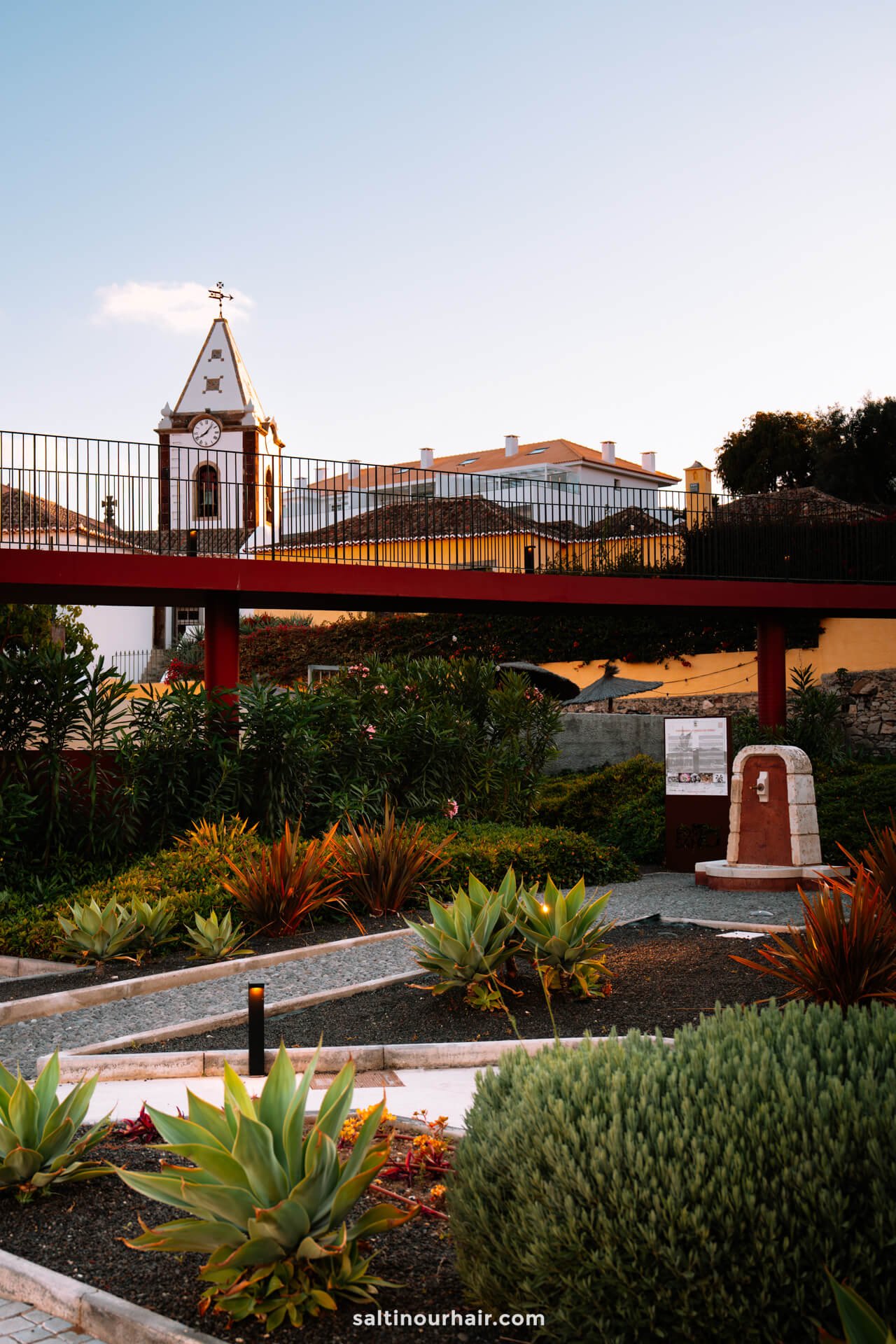 A landscaped garden with various plants, a small monument, and a raised pedestrian walkway; in the background, the white church tower with a cross rises above Vila Baleira, Porto Santo, Madeira.