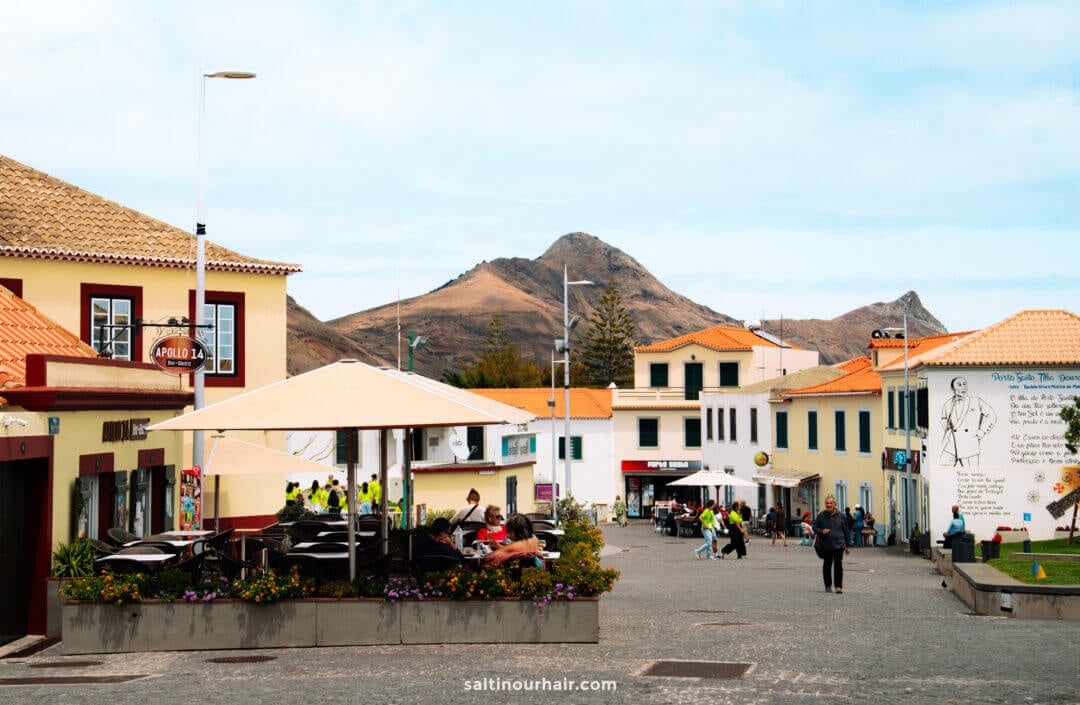 A lively Porto Santo town square with outdoor seating, people walking, colorful buildings, and a mountain in the background under a partly cloudy sky