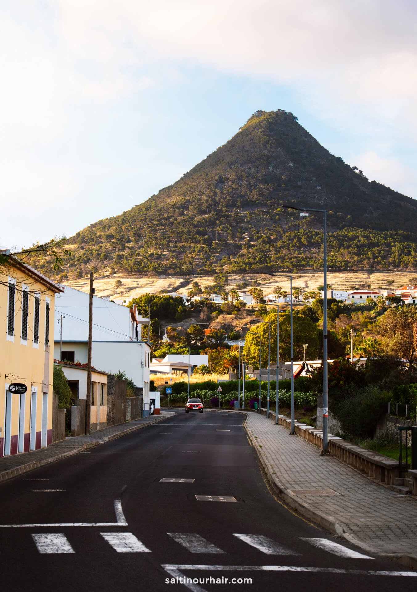 A quiet, empty street in Porto Santo leads toward a conical, green hill under a partly cloudy sky, with houses and trees lining the roadsideâ€”hinting at peaceful moments before exploring the many things to do in Porto Santo.