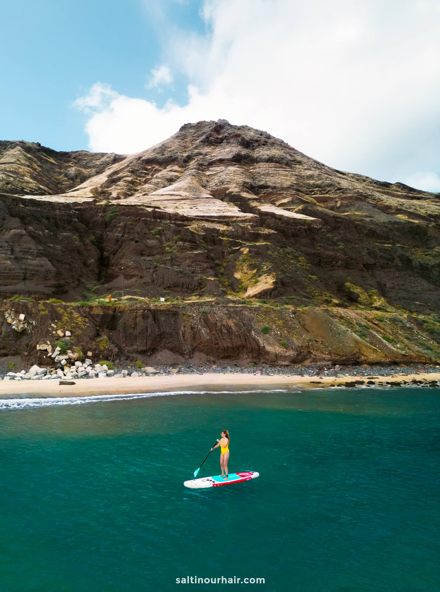 A woman paddleboarding on clear blue water near a rocky, mountainous shoreline under a partly cloudy skyâ€”one of the unforgettable things to do in Porto Santo.