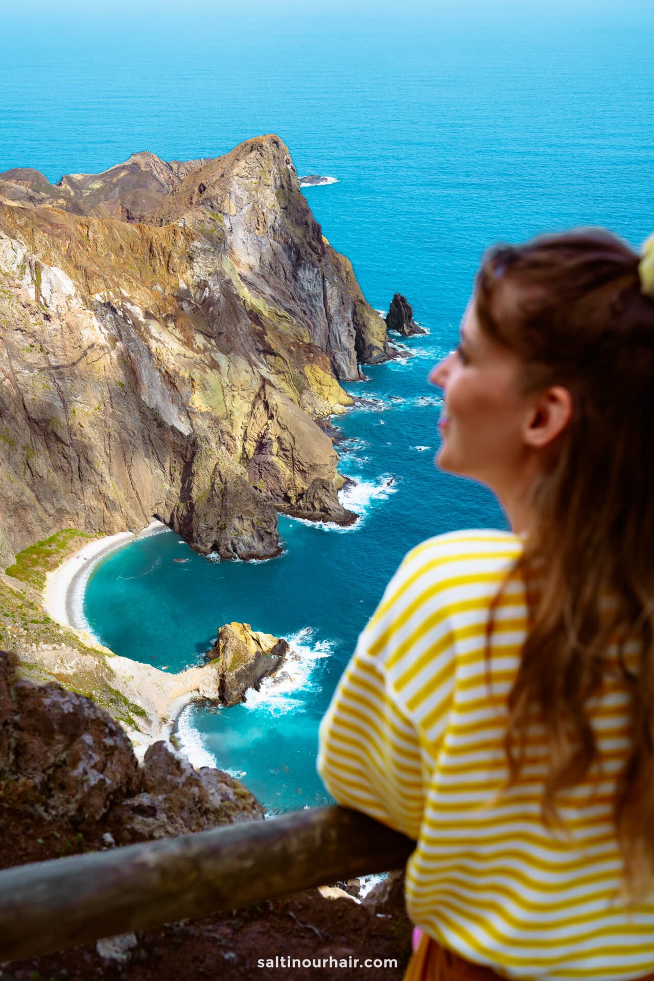 A woman in a yellow striped shirt looks out over Porto Santoâ€™s rocky coastline with turquoise water and steep cliffs, capturing the beauty of one of the top things to do in Porto Santo.