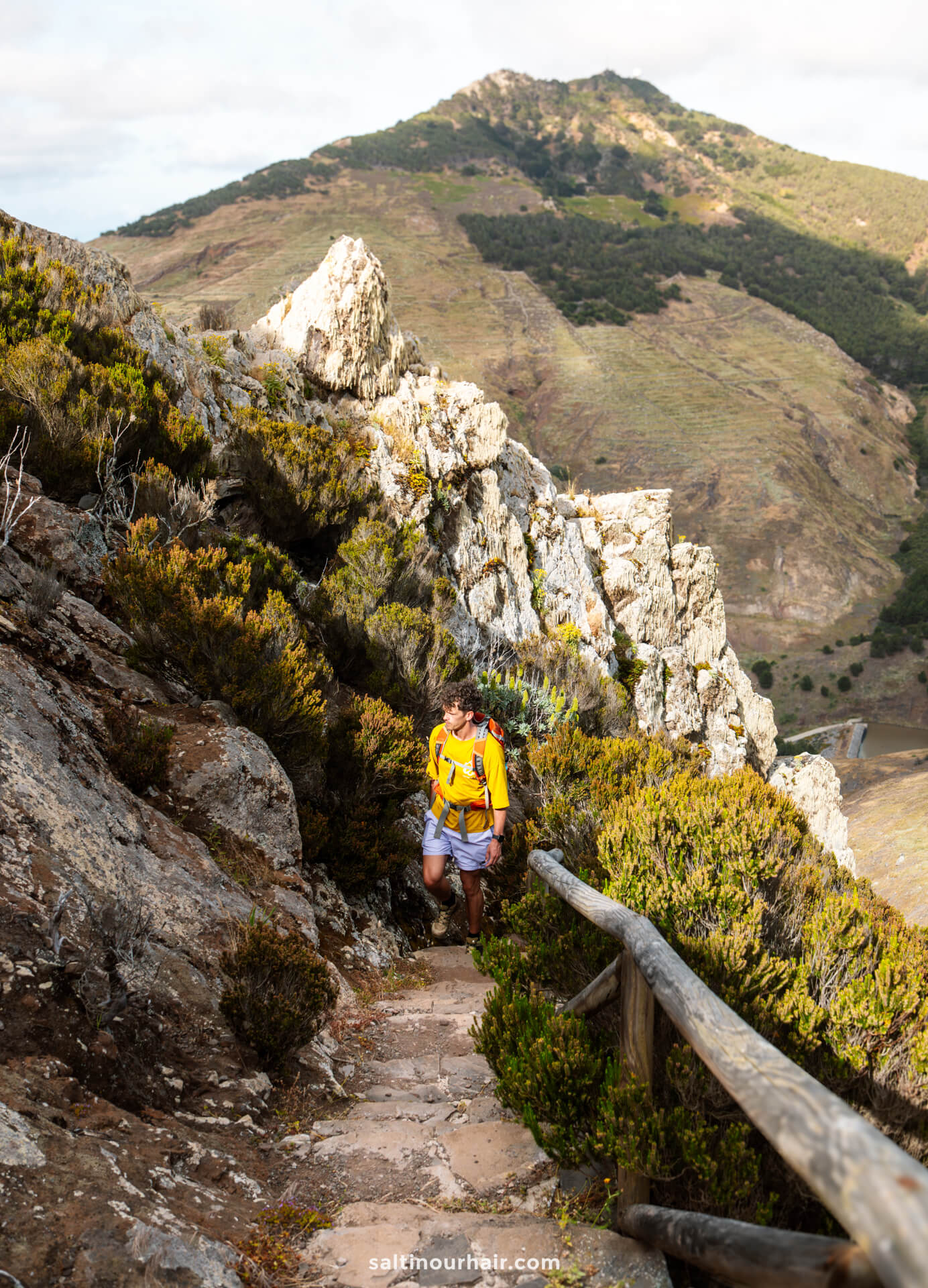 A man wearing a yellow backpack hikes up a narrow rocky trail with a wooden railing, surrounded by mountains and shrubsâ€”a perfect scene among the many things to do in Porto Santo.
