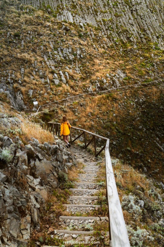 A woman in a yellow jacket walks up a wooden and stone path with a railing along a rocky, grassy hillsideâ€”one of the scenic things to do in Porto Santo.