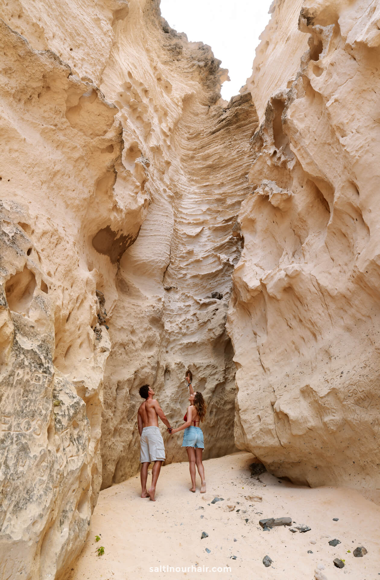 a couple stand at the base of a tall, narrow rock canyon with textured sandstone walls, looking upwardâ€”an adventure reminiscent of exploring hidden gems among the things to do in Porto Santo.