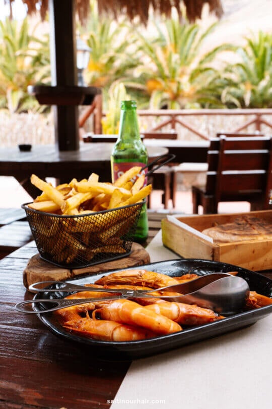 A plate of cooked shrimp, a basket of French fries, and a bottled drink on a wooden table at an outdoor dining area with palm trees in the backgroundâ€”perfect for enjoying while exploring things to do in Porto Santo.