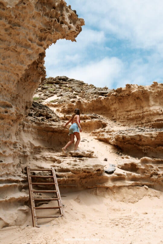 A person in a tank top and shorts climbs natural rock steps near a wooden ladder, surrounded by rugged sandstone formations under a partly cloudy skyâ€”one of the adventurous things to do in Porto Santo.