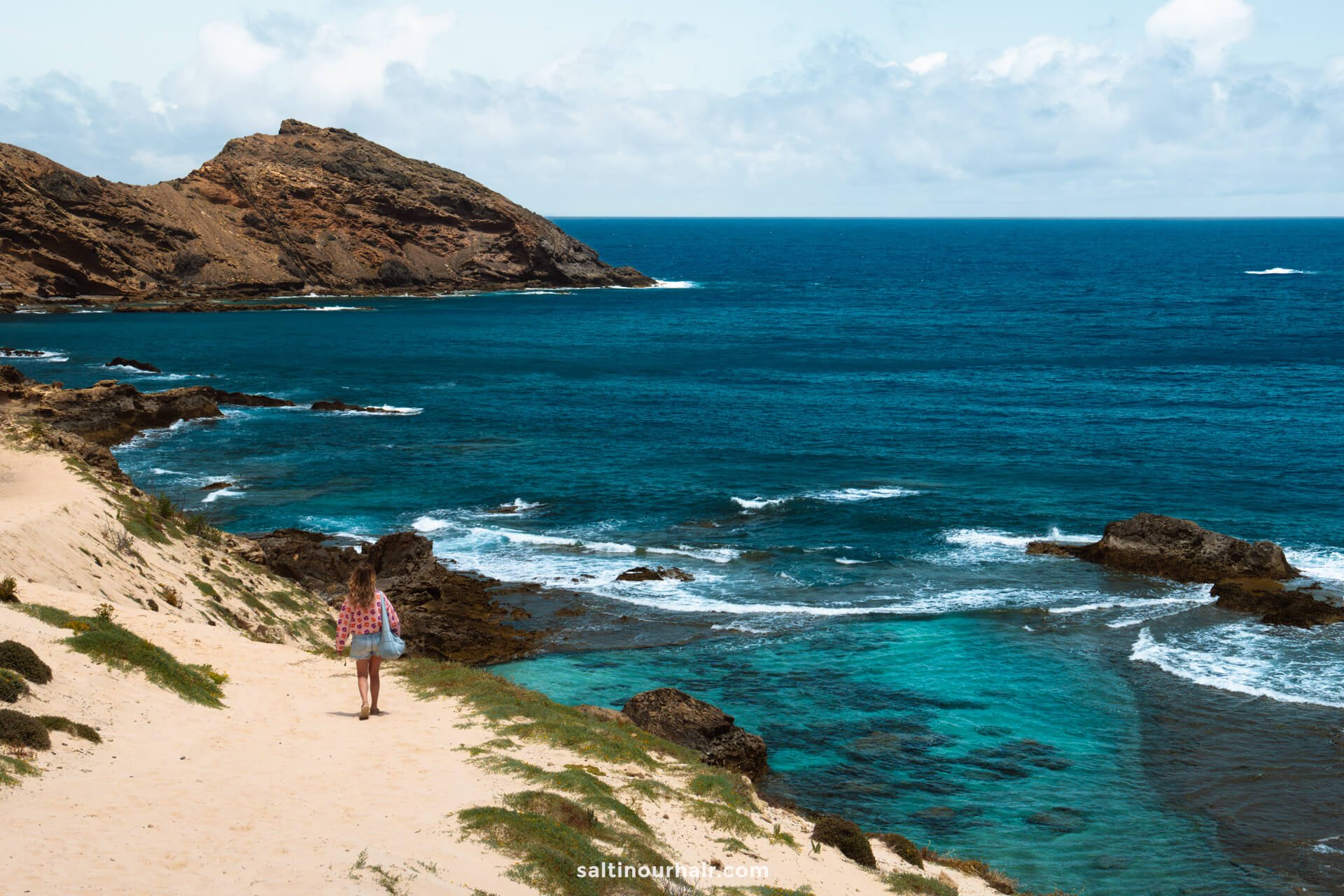 A woman walks along a sandy path by the rocky Porto Santo coastline, with clear blue ocean water and a large hill in the backgroundâ€”capturing just one of the many scenic things to do in Porto Santo.