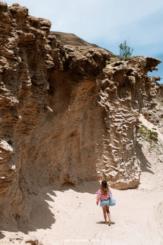 A woman with long hair walks along a sandy path beside large, rugged rock formations under a clear skyâ€”one of the scenic things to do in Porto Santo.