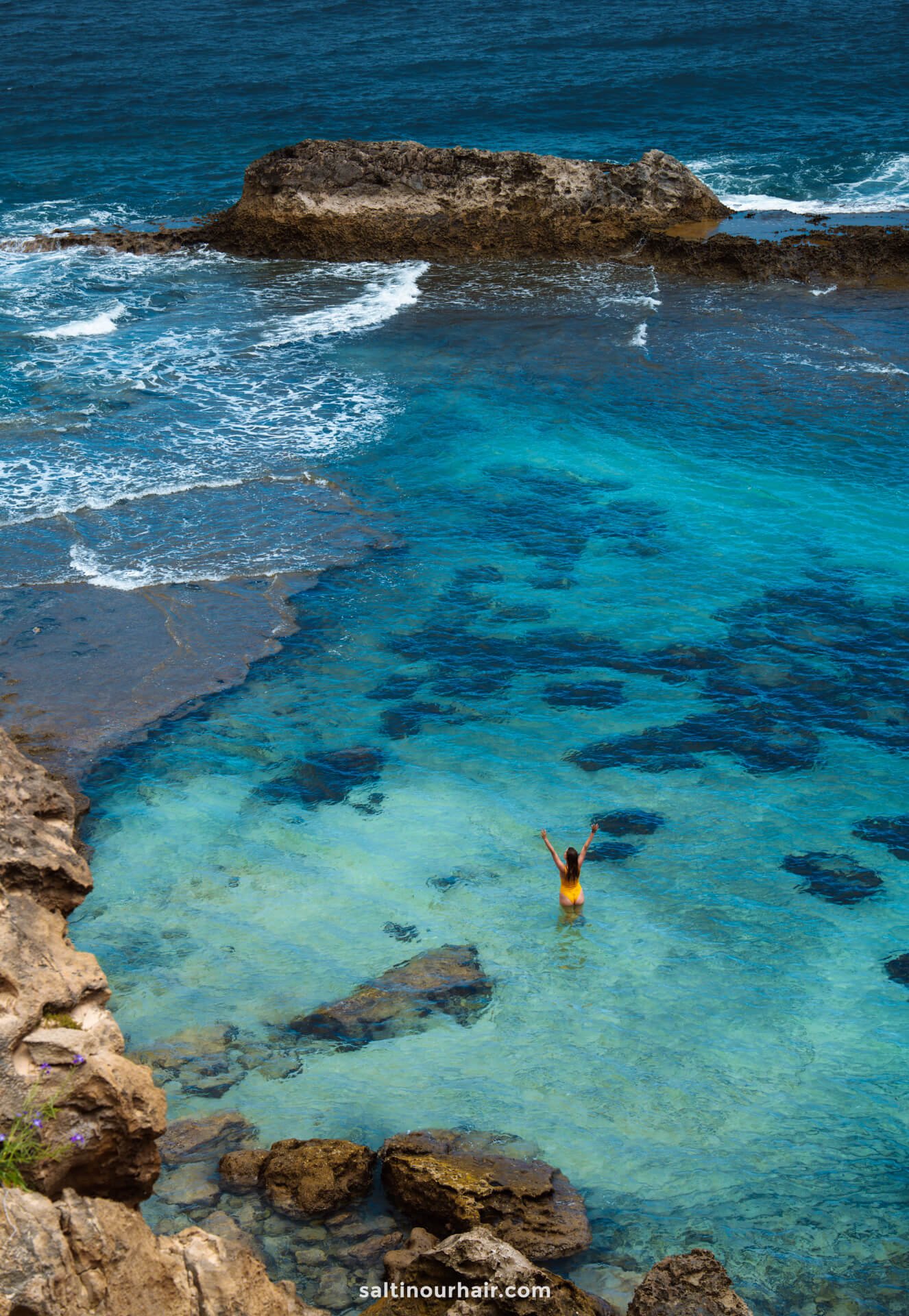 A woman in an yellow swimsuit stands in a natural pool in porto santo madeira near rocky cliffs by the ocean, raising their arms toward the sky