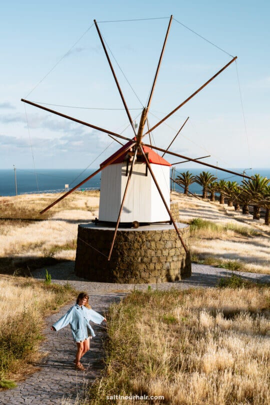 A girl in a blue dress walks on a stone path near a large traditional windmill in a grassy field by the sea—one of the charming things to do in Porto Santo, madeira