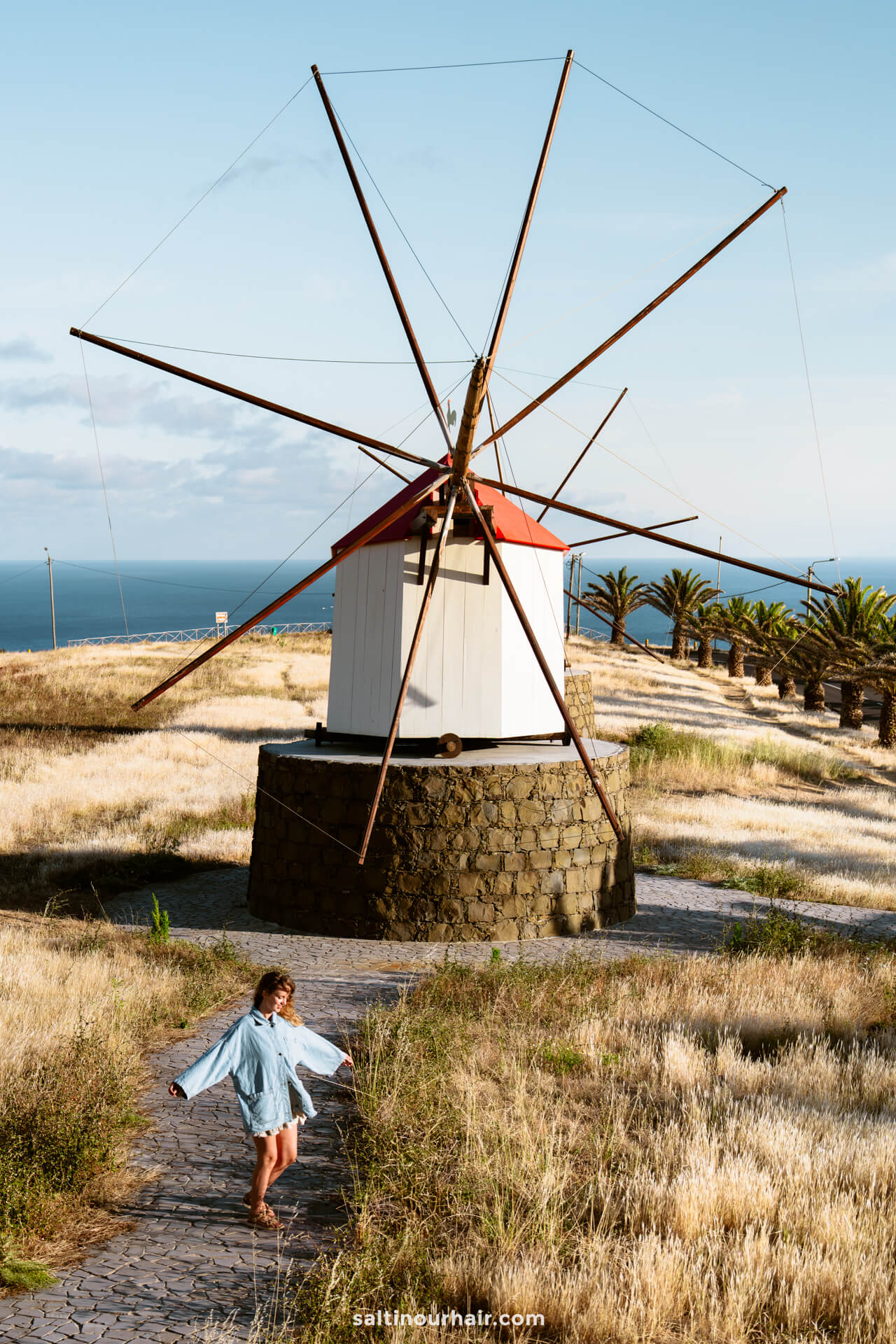 A girl in a blue jacket walks on a stone path near a large traditional windmill in a grassy field by the seaâ€”one of the charming things to do in Porto Santo.