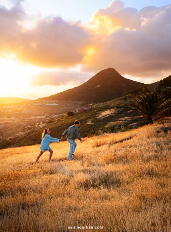 Two people walk hand in hand through a golden grassy field at sunset on Porto Santo, with a mountain and scattered clouds in the backgroundâ€”a perfect scene among the many things to do in Porto Santo.