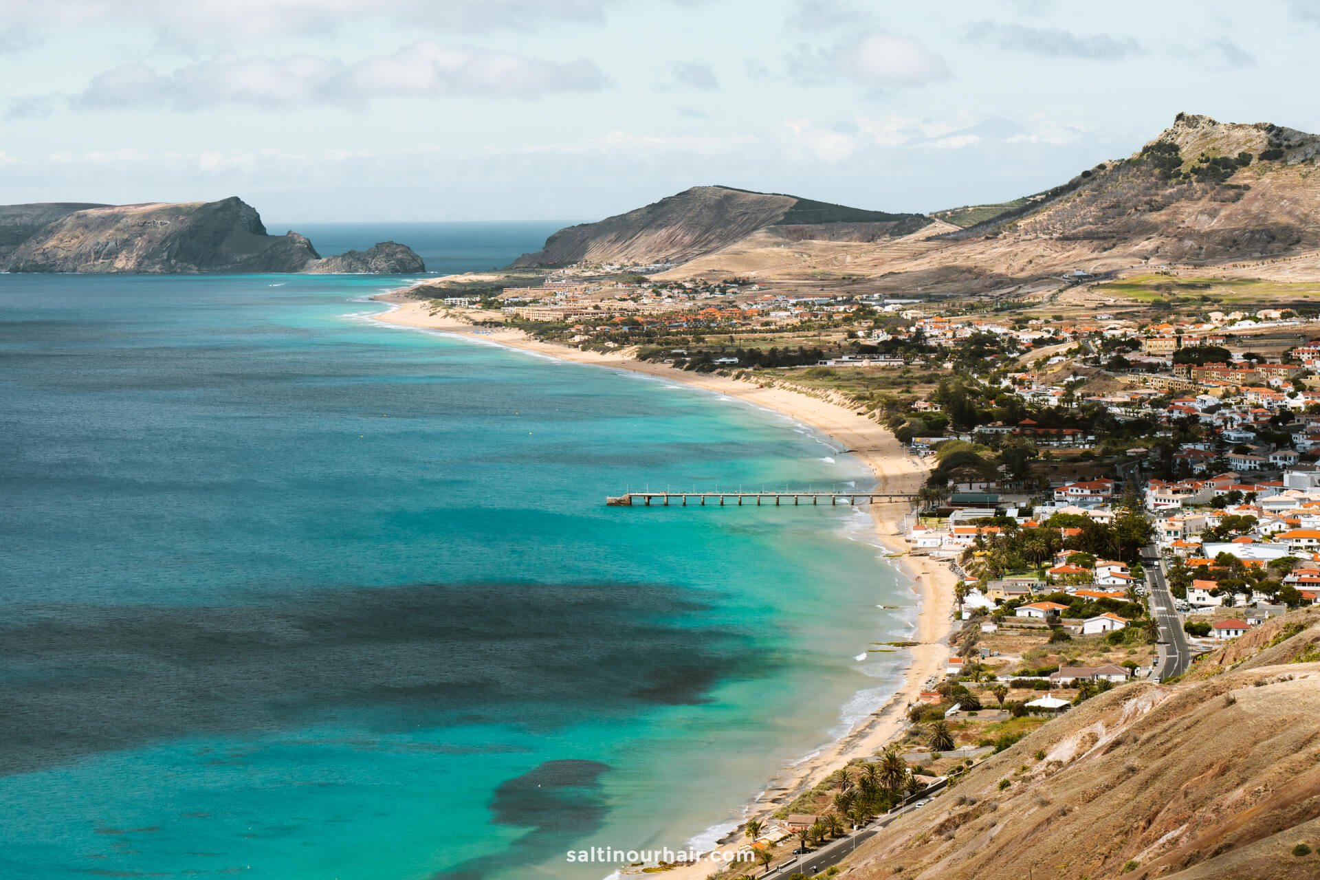 coastal town with white buildings lines a sandy beach, turquoise waters, and a long pier as seen from the breathtaking viewpoint Porto Santo, bordered by green hills and rugged mountains under a partly cloudy sky.