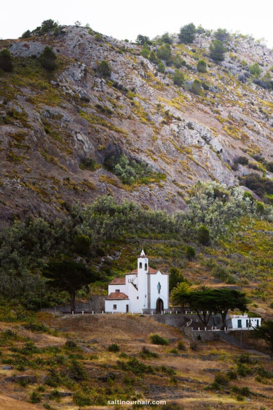 A small white church stands at the base of a large, rugged hillside with sparse vegetation and some trees nearbyâ€”a tranquil spot reflecting the natural beauty of Porto Santo and offering a peaceful escape among the things to do in Porto Santo.