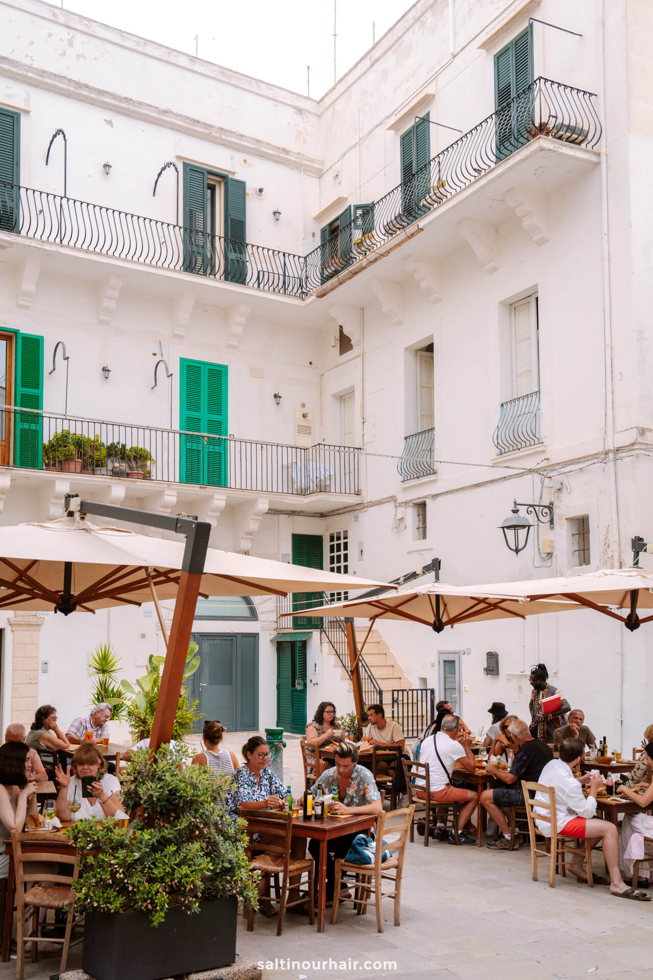 People sit at tables under umbrellas in a sunny courtyard in Monopoli Italy.
