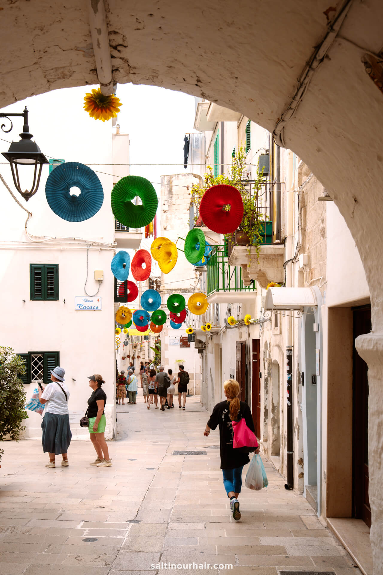 A narrow street in Monopoli, Italy is decorated with colorful paper lanterns. People are walking along the street under an arched passageway.