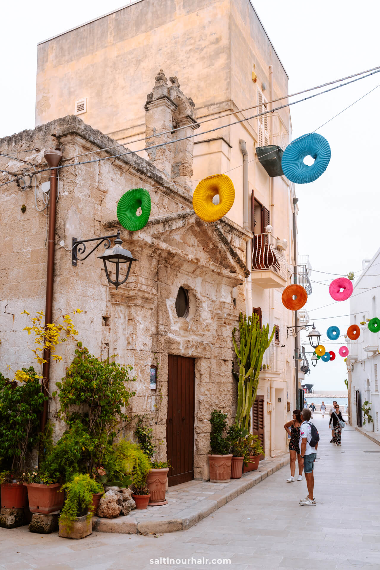 A narrow street in Monopoli, puglia Italy, with stone buildings, potted plants, colorful circular decorations hanging overhead, and a few people walking.