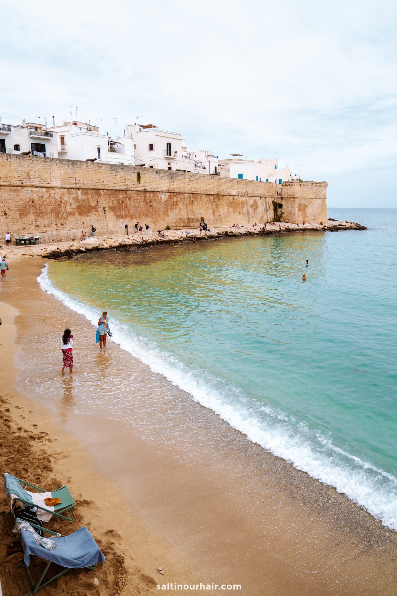 People stand and swim along a sandy beach of cala porta vecchia with calm waves in Monopoli, puglia Italy, bordered by a stone wall and white buildings above