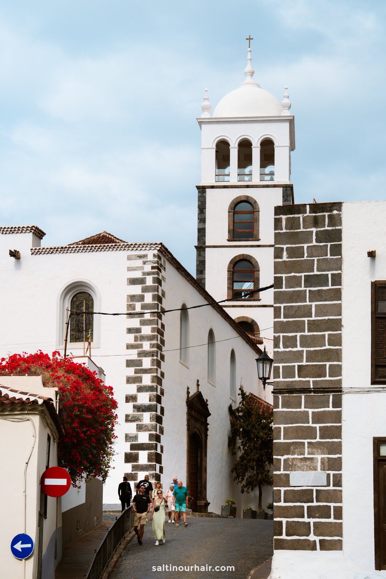 White church with a tall bell tower, stone accents, and arched windows stands on a narrow cobblestone street in Garachico, Tenerife, with people walking and flowers on the left.